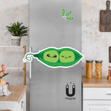 Product picture of a cute flexible two peas in a pod fridge magnet with a background example of pea pod magnets clinging to a stainless steel fridge. Peas in a pod magnet is designed by Bare It Designs.