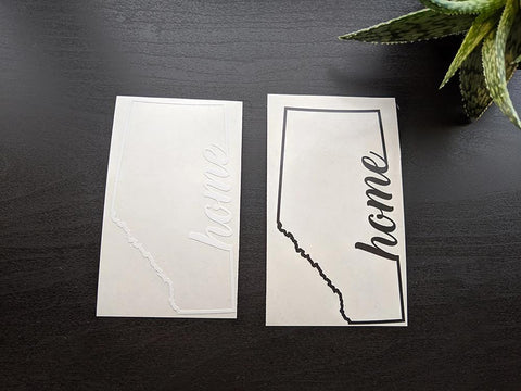 White and black vinyl sticker/decal with the outline of the province of Alberta and the word home in script font imbedded on the right side of the outline. Created by Bare It Designs, Edmonton, AB, Canada.
